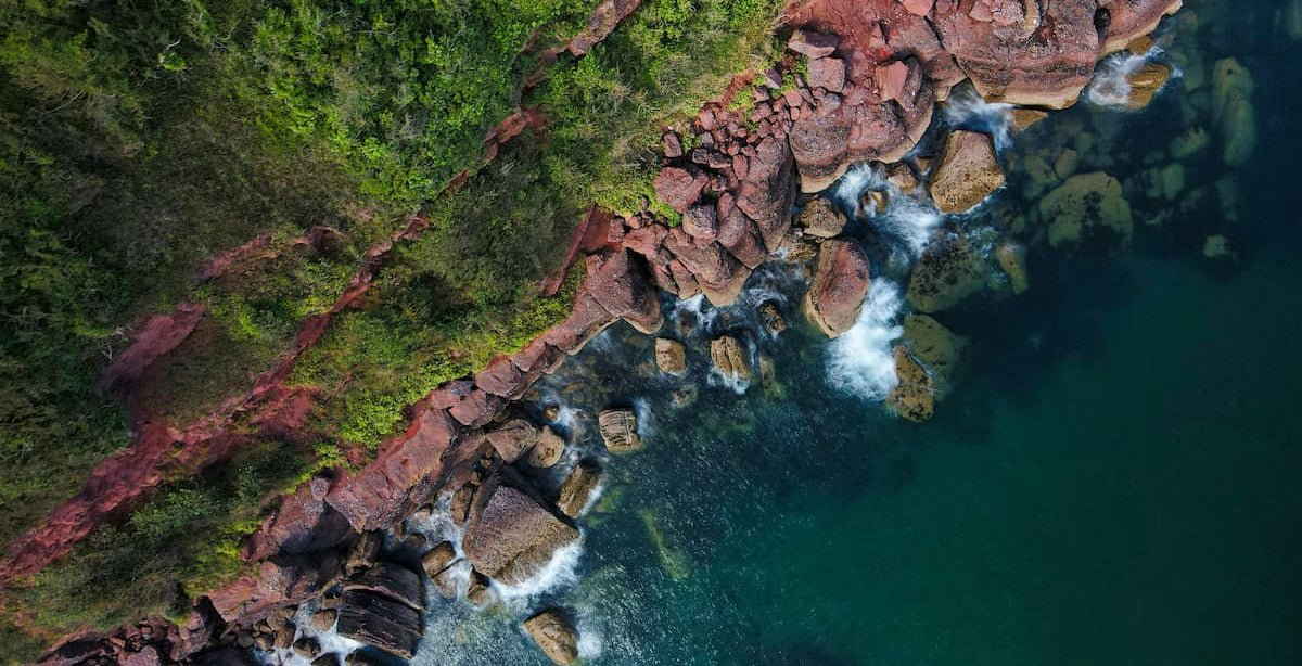 Aerial view of red rocks meeting a turquoise sea below green coastal forest