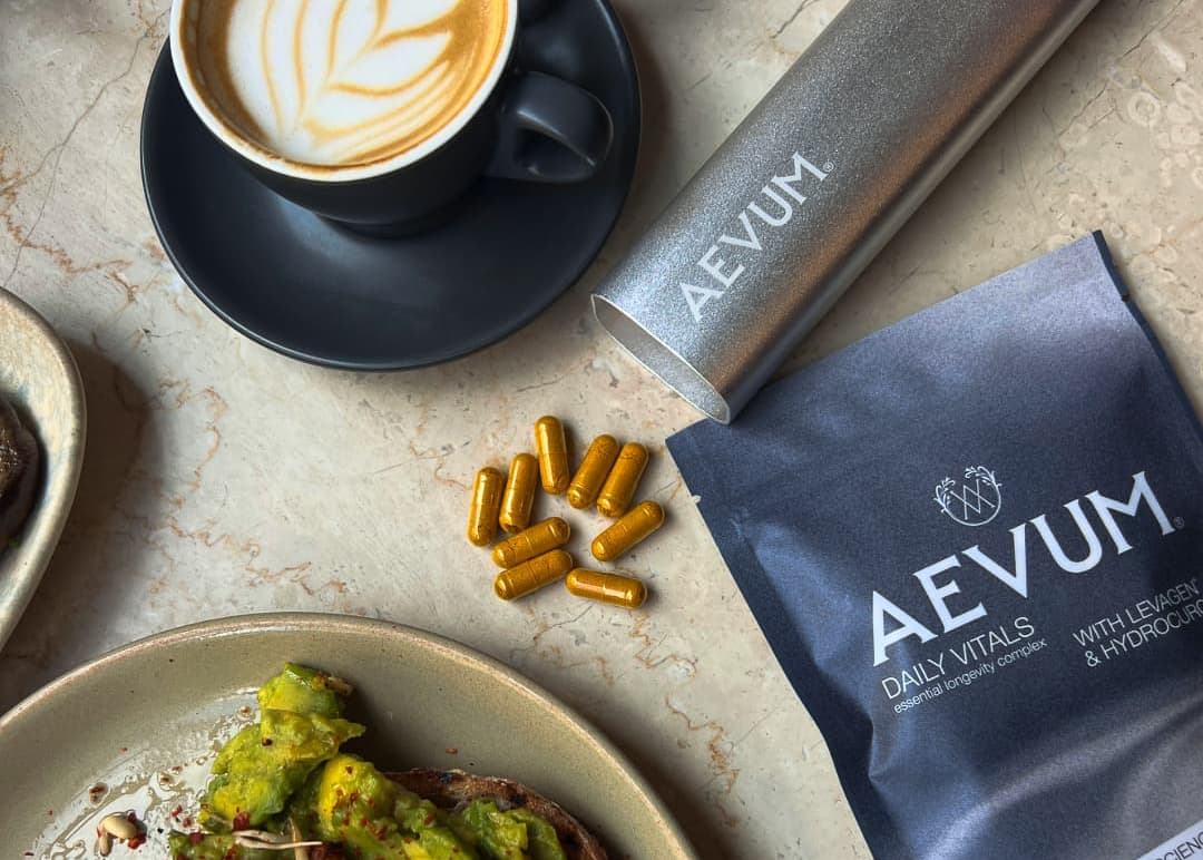 A styled flat-lay of AEVUM Daily Vitals capsules, showing them with AEVUM's aluminium pill case and the dark grey Daily Vitals pouch. Around the capsules is a black coffee cup and plate of avocado toast on a marble surface.