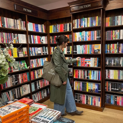 A woman in a green trench coat and blue jeans browses bookshelves inside a cozy bookstore, carrying an AEVUM tote bag. The shelves are filled with books under dark wood signs labeled 'FICTION'.