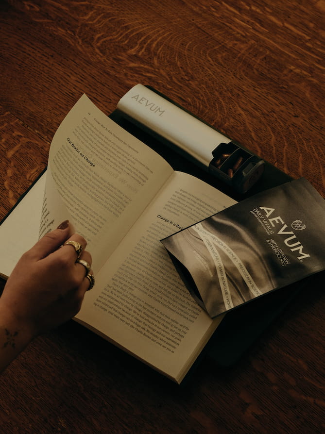Hand turning the page of a book beside a Daily Vitals pouch and AEVUM aluminium pill case on a wooden table