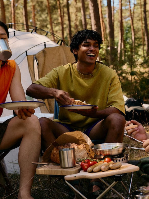 Friends sharing a meal and laughing at a campsite in a pine forest, illustrating AEVUM's energy production pillar.